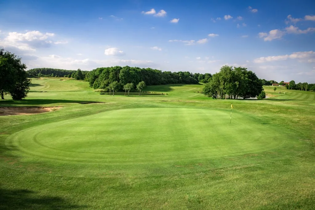 A lush green golf course in bristol under a bright blue sky with scattered white clouds, featuring a manicured putting green in the foreground and rolling hills with trees in the background