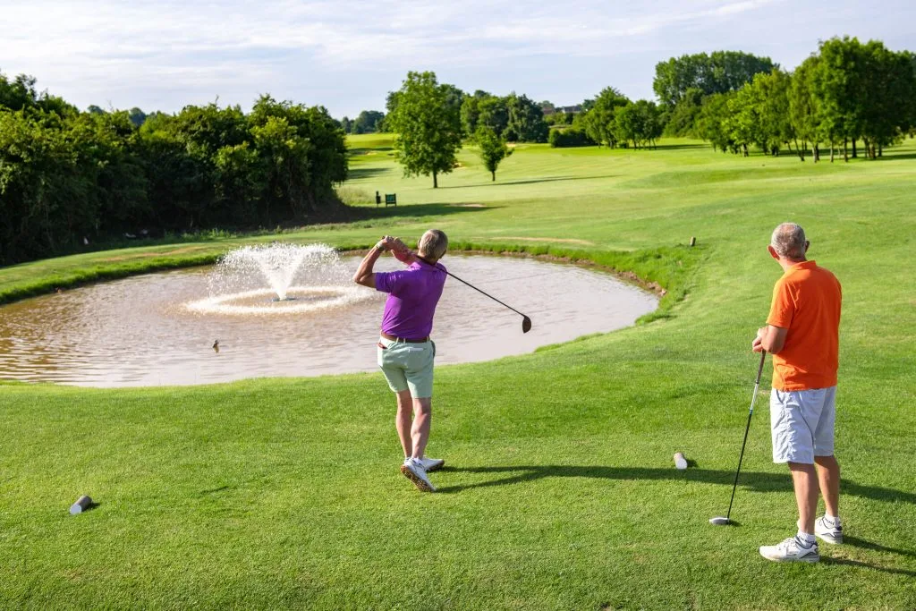 Two men hitting golf shots over a water fountain at a golf course in Bristol.